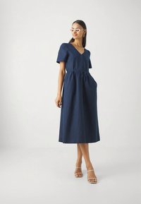 Ženska oblečena v temno modro midi obleko s kratkimi rokavi in gumbom, bele sončne očala, zlate uhane v obliki obroča in bež sandale s trakovi, stoji na beli podlagi.