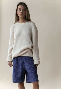 Cremfarbener Strickpullover mit strukturiertem Rippenmuster, kombiniert mit navyblauen Shorts mit weißen Punkten. Der Pullover hat umgeschlagene Ärmel und eine lockere Passform.