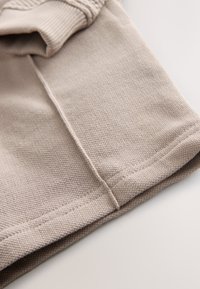 Tela beige claro con superficie texturizada, que presenta un puño acanalado y costuras planas. El dobladillo está cuidadosamente terminado.