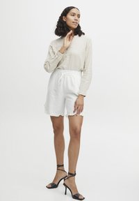 Shorts en lin blanc avec un détail de ourlet volants, associés à une chemise beige à bouton et des sandales à talons hauts noires à lanières.