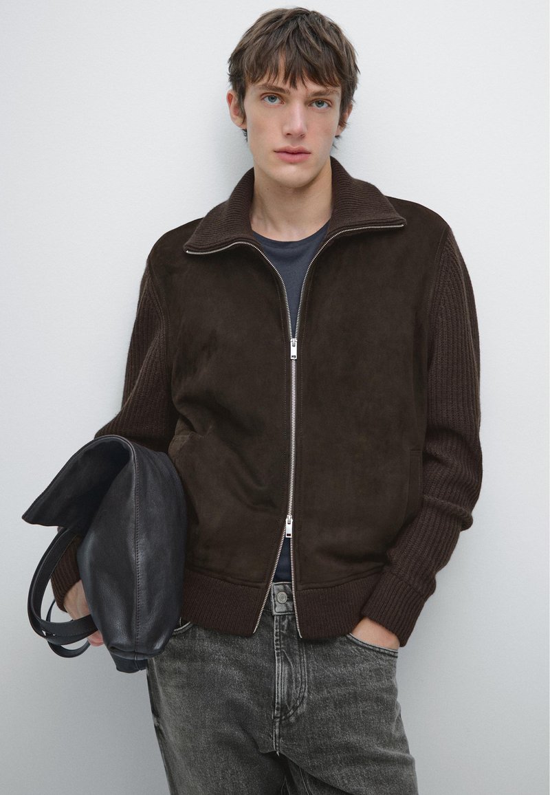 Brązowa skórzana kurtka z zamszu, z ribbowymi rękawami, zamek błyskawiczny z przodu i bocznymi kieszeniami. Model trzyma czarną skórzaną torbę, w połączeniu z szarymi dżinsami.