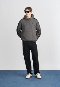 Sweatshirt cinza com capuz, com um bolso frontal e logotipo preto, combinado com calças pretas e ténis brancos. O fundo é cinza-claro.