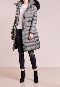 Světlý zelený péřový kabát s kapucí lemovanou kožešinou, doplněný krémovým svetr s copánkovým vzorem a černými roztrhanými džíny. Obuv tvoří černé kotníkové boty na podpatku.