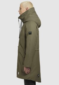 Parka verde oliva con colletto alto, cordini regolabili e ampie tasche frontali. Presenta una finitura testurizzata e una patch con logo sulla manica.