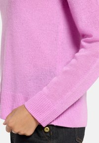 Pull en tricot rose clair avec poignets et ourlet côtelés, doté d'une texture douce et d'une coupe ajustée. Détail de couture visible sur la manche.