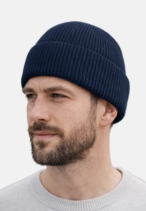 Bearded man wearing a navy blue ribbed knit beanie and a light grey crew neck sweater, looking slightly left on a plain white background.