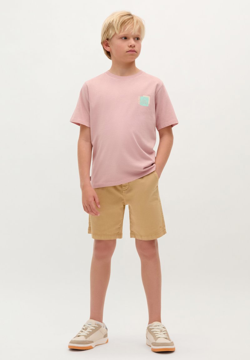 Vaaleanpunainen lyhythihainen t-paita, jossa on mintunvihreä logopatch, yhdistettynä khaki-lyhyisiin housuihin. Beige lenkkarit, joissa on valkoiset nauhat ja oranssit yksityiskohdat.