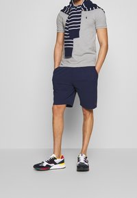 Man som bär en grå pikétröja med marinblå shorts, en randig marinblå tröja över axlarna och flerfärgade sportskor, med händerna i fickorna.