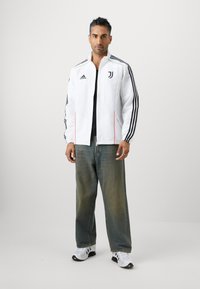 Hvid track jakke med sorte striber; har røde detaljer og Juventus logo. Bæres med løse denim jeans og hvide sportssko.