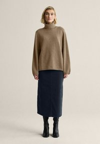 Ruskea kauluspaidan tyyli, jossa on reilu istuvuus, yhdistetty tummansinisään polvipituiseen hameeseen ja mustiin nilkkureihin neutraalilla taustalla.