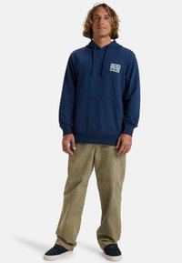 Sudadera azul marino con cordón, bolsillo tipo canguro y pequeño parche con logo de ola. Combinada con pantalones de pana verde claro y zapatos oscuros.