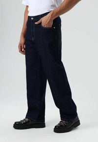 Donkerblauwe spijkerbroek met een klassieke rechte pijp, voorzien van gele stiksels en voorzakken, gecombineerd met zwarte veterlaarzen.