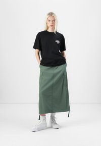 Black cotton t-shirt with a colorful logo on the chest, paired with a green skirt featuring cargo pockets and white sneakers.
