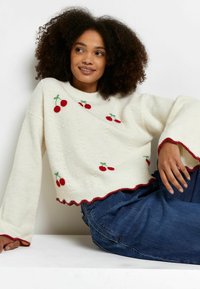 Krämfärgad stickad tröja med röd körsbärsbroderi, scalloped nederkant och vida ärmar. Kombineras med blåjeans, visad mot en neutral bakgrund.