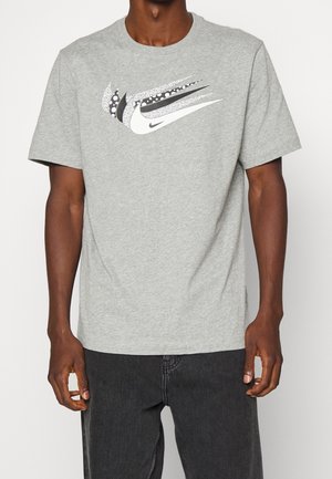 Mies pukeutunut vaaleanharmaaseen lyhythihaiseen t-paitaan, jossa on monikuvioinen mustavalkoinen Nike swoosh -logo, yhdistettynä mustiin farkkuihin.