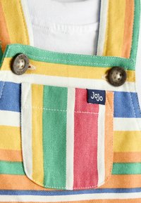 Krāsainas svītrainas kombinezons ar priekšējo kabatu, brūnām pogām, uz kurām iegravēts "JoJo", nēsāts virs baltas krekla.