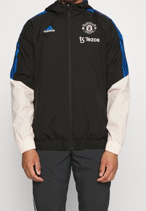 Czarna kurtka Manchester United na zamek z niebieskimi paskami na ramionach, białymi rękawami, logo Adidas i brandingiem sponsora Tezos.