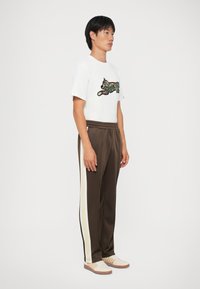 Witte t-shirt met een camouflagekat-graphic, gecombineerd met bruine trainingsbroek met contrasterende witte zijkanten en een elastische tailleband.