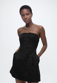 Γυναίκα με κοντά μαλλιά που φοράει έναν μαύρο αμάνικο φόρεμα με υφή και σχέδια με λουλούδια, στέκεται με τα χέρια στις τσέπες μπροστά σε ένα απλό φόντο.