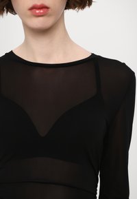 Juodas ilgomis rankovėmis viršus, pagamintas iš skaidraus audinio, su V formos iškirpte ir prigludusia konstrukcija, kuri pabrėžia apatinių drabužių detales.