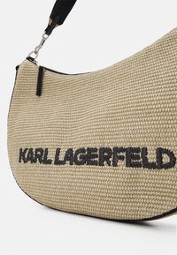 Sac à épaule en tissu tissé beige avec une sangle noire et "KARL LAGERFELD" brodé en noir sur la face avant.