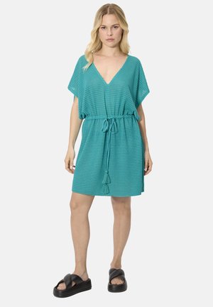 Mujer con vestido texturizado de pico en color verde azulado, de manga corta y cinturón en la cintura, combinando con sandalias negras de plataforma tipo deslizador, posando sobre fondo blanco.