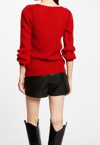 Rode gebreide trui met een gestructureerde afwerking, voorzien van pofmouwen en een brede halslijn, gecombineerd met zwarte leren shorts en hoge zwarte laarzen.
