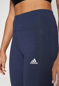 Tamsiai mėlynos sportinės leggingas su aukštu liemeniu, su balta Adidas logotipu kairiajame klube. Lygi, elastinga medžiaga be raštų.