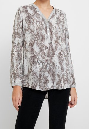 Femme portant une chemise henley à manches longues boutonnée avec un motif serpent gris et marron, associée à un pantalon noir.