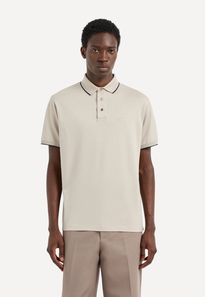 Polo de color beige claro con cuello y tapeta de tres botones, presenta ribete negro a lo largo del cuello y las mangas. Textura suave.