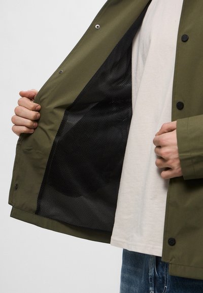 Giacca verde oliva con chiusura a bottone, dotata di una fodera in rete nera traspirante. Il materiale esterno sembra liscio e resistente.