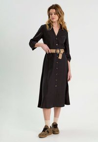 Robe midi noire boutonnée avec manches longues et deux poches poitrine, accentuée par une ceinture tressée beige. Associée à des baskets à imprimé léopard.