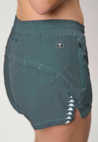 Prim-plan lateral cu o persoană purtând pantaloni scurți sport de culoare verde închis, cu un buzunar cu fermoar și un model de logo alb lângă tiv.