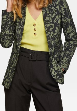 Vrouw draagt een blazer met groen bladmotief, een geel geribd topje met drie knopen en zwarte hoge taille broek met brede riem.