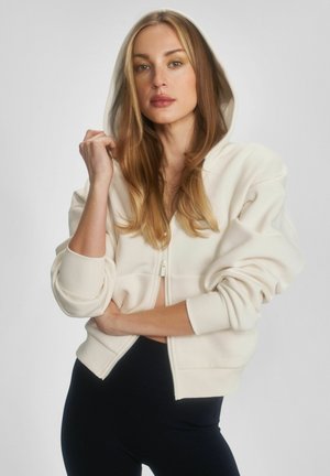 Kobieta o długich blond włosach, ubrana w kremową bluzę z kapturem na suwak, z założonym kapturem oraz czarne spodnie, stojąca na tle jednolitego tła.