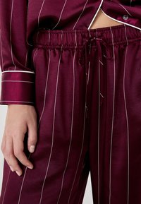 Burgundyröd satin pyjamaset med vita vertikala ränder. Slät textur, elastisk midja med dragsko och kantade ärmslut.