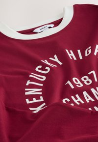 Tummanpunainen puuvillainen t-paita valkoisella pyöreällä pääntiellä. Suuri, valkoinen, pyöreä tekstikuvio, jossa lukee "Kentucky High". Pehmeä tuntu.