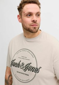 Beige bomulds-T-shirt med et rundt sort grafisk tryk med tekst og diamantmønster. Kortærmet og med rund hals.