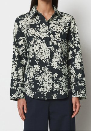 Chemise noire boutonnée avec motif floral blanc, portée avec un pantalon foncé, montrant le haut du corps et les bras sur fond uni.