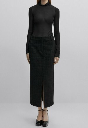Femme portant un col roulé noir à manches longues, une jupe texturée foncée taille haute avec une fente devant, et des talons compensés noirs à bride à la cheville.