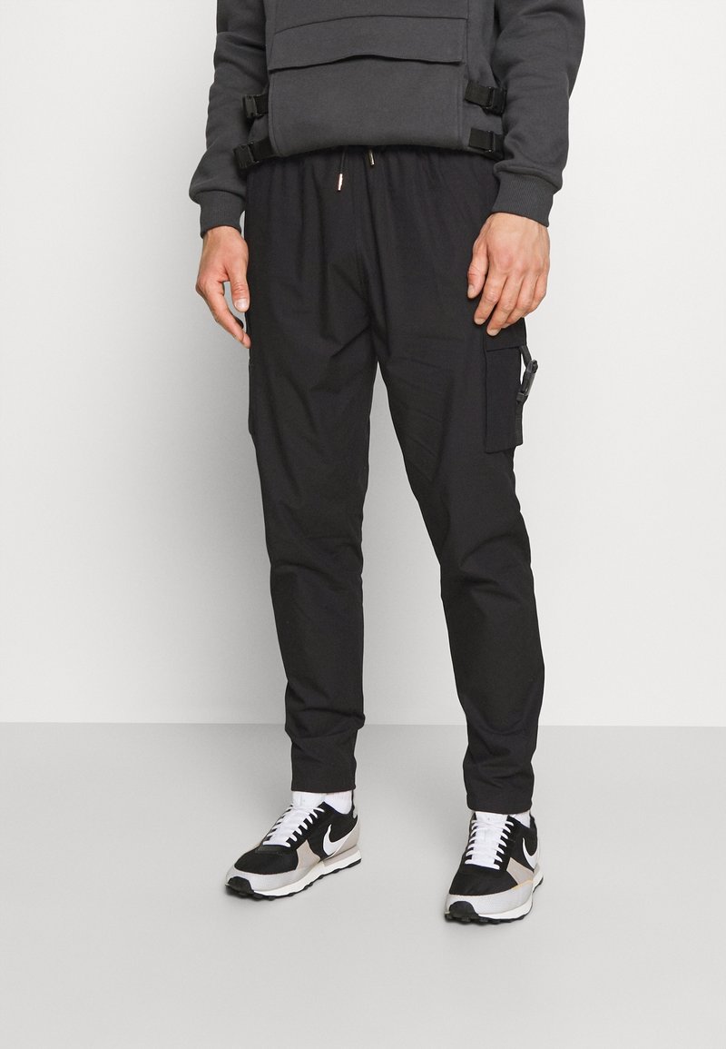 Homme portant un pantalon cargo noir avec poche latérale et des baskets Nike noires et blanches, debout devant un fond clair uni.