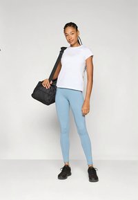 Baltas trumpomis rankovėmis marškinėliai, šviesiai mėlynos tamprės, juodi sportbačiai ir juoda sportinė rankinė su petneša. Modelis stovi priešais šviesų foną.