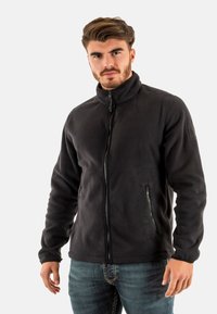 Chaqueta de forro polar negra con un cuello alto, cierre de cremallera frontal y bolsillos laterales. Textura suave, ajuste casual y sin patrones o herrajes visibles.