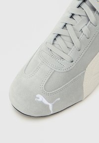Γκρι σουέτ αθλητικά παπούτσια με λευκό λογότυπο Puma, υφασμάτινη επιφάνεια, στρογγυλή μύτη και επίπεδα κορδόνια. Πλευρική λεπτομέρεια σε ανοιχτό μπεζ τόνο.