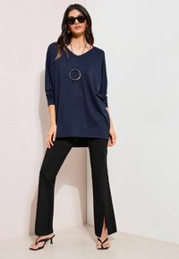 Teget dugi rukav s labavim krojem, uparen s crnim hlačama trapeznog kroja koje imaju bočne proreze, te crnim sandalama s remenčićima. Naočale za sunce i okrugla ogrlica.