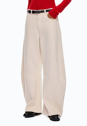 Persona che indossa pantaloni beige a gamba larga, cintura nera con fibbia decorativa, scarpe nere e maglia rossa a manica lunga con la mano in tasca.