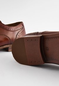 Chaussures en cuir marron présentant un détail de brogue complexe, une semelle texturée et un talon empilé avec un motif de bande de roulement subtil. Taille indiquée : 42.