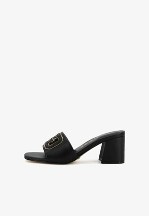 Sandalo con tacco a molla nero realizzato in materiale sintetico liscio. Caratterizzato da una larga fascia con un logo in oro e un tacco a blocco spesso.