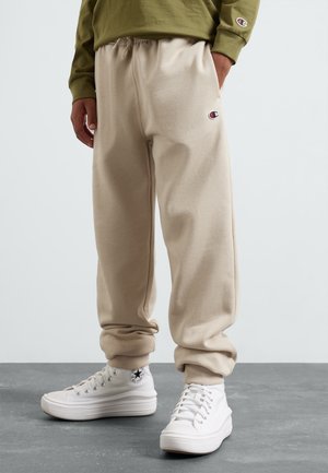 Person iført beige joggingbukser og hvide høje sneakers står på gråt gulv, grøn langærmet trøje synlig.