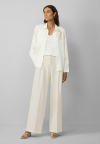 Crème zijden blouse met bloemenbroderie, gecombineerd met licht crème wijde broek en witte sandaaltjes met bandjes. Ontspannen pasvorm met knoopsluiting.